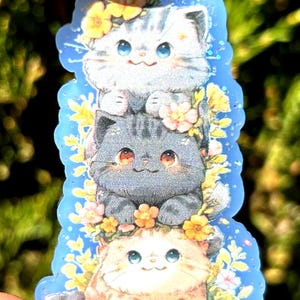 May include: A sticker featuring three stacked cartoon cats with floral accents. The top cat is gray and white, the middle cat is gray, and the bottom cat is tan. Each cat has large blue eyes and is surrounded by yellow and pink flowers.