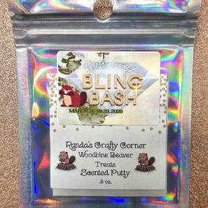 Könnte beinhalten: Eine holografische Tüte mit einer kleineren Tüte mit dem Text "West Coast Bling Bash" und "March 26 to 29, 2026". Die kleinere Tüte enthält auch "Randa's Crafty Corner Woodbine Beaver Treats Scented Putty 85g."