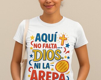 May include: White t-shirt with the text "AQUÍ NO FALTA... DIOS NI LA AREPA" in blue, yellow, and red. The design includes a cross, stars, and a stylized arepa. The shirt is worn by a person.