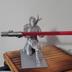 May include: A silver 3D printed figurine of a Star Wars character holding a red lightsaber. The character is kneeling and wearing a helmet and armor. The figurine is on a gray base.