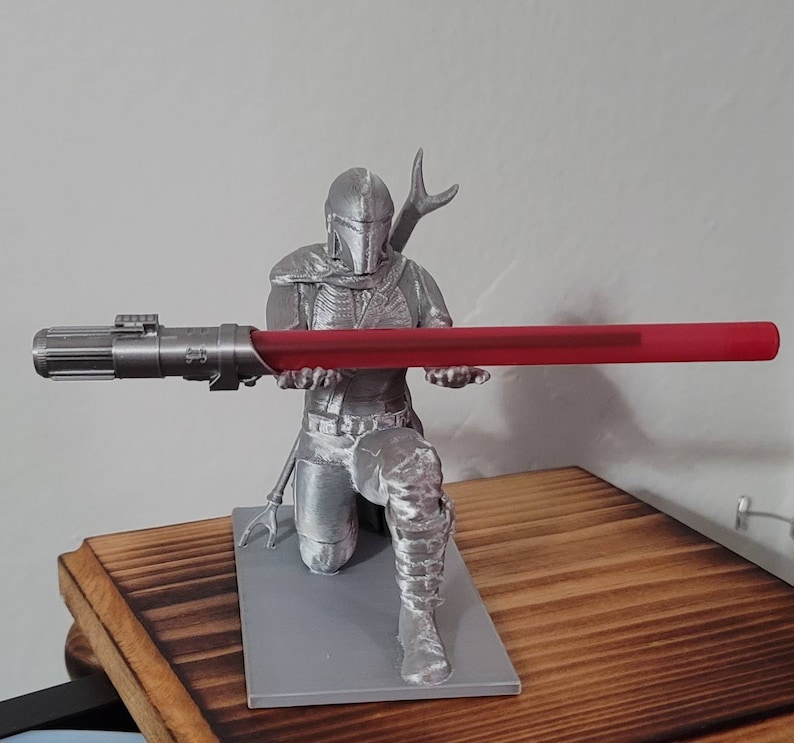 May include: A silver 3D printed figurine of a Star Wars character holding a red lightsaber. The character is kneeling and wearing a helmet and armor. The figurine is on a gray base.
