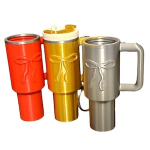 Könnte beinhalten: Drei Reisebecher in verschiedenen Farben mit einem Schleifendesign. Die Becher sind rot, gold und silber. Der silberne Becher hat einen Griff.
