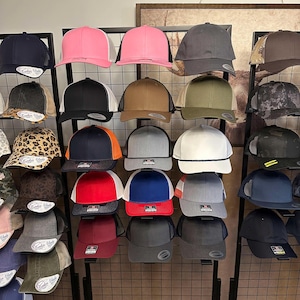 Könnte beinhalten: Eine Präsentation verschiedener Baseballkappen in verschiedenen Farben, darunter Rosa, Schwarz, Grau und Braun. Einige Kappen haben Mesh-Rücken und Seildetails. Die Kappen sind auf einem schwarzen Metallgitter angeordnet.