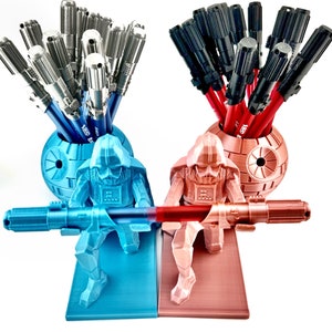 May include: Two Star Wars themed pencil holders, one blue and one red, each featuring a Darth Vader figurine holding a lightsaber. The blue holder has a Death Star design and holds silver pens, while the red holder has a Death Star design and holds black pens.