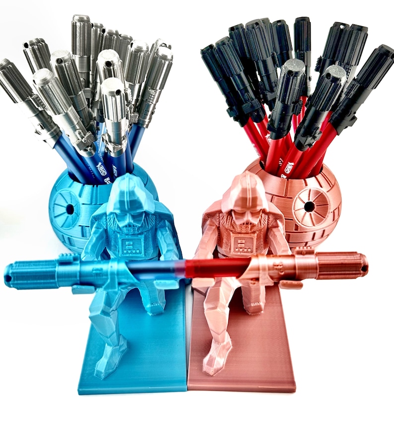 May include: Two Star Wars themed pencil holders, one blue and one red, each featuring a Darth Vader figurine holding a lightsaber. The blue holder has a Death Star design and holds silver pens, while the red holder has a Death Star design and holds black pens.