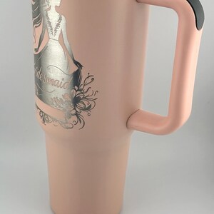 May include: A pink stainless steel tumbler with a black handle and a silver engraved design of a woman in a wedding dress with the word "bridesmaid".