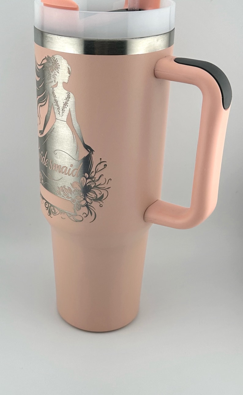 May include: A pink stainless steel tumbler with a black handle and a silver engraved design of a woman in a wedding dress with the word "bridesmaid".