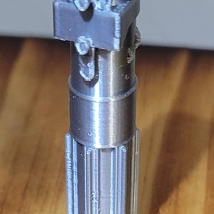 May include: A silver-colored 3D printed lightsaber hilt with a detailed design. The hilt has a textured surface and a cylindrical shape.