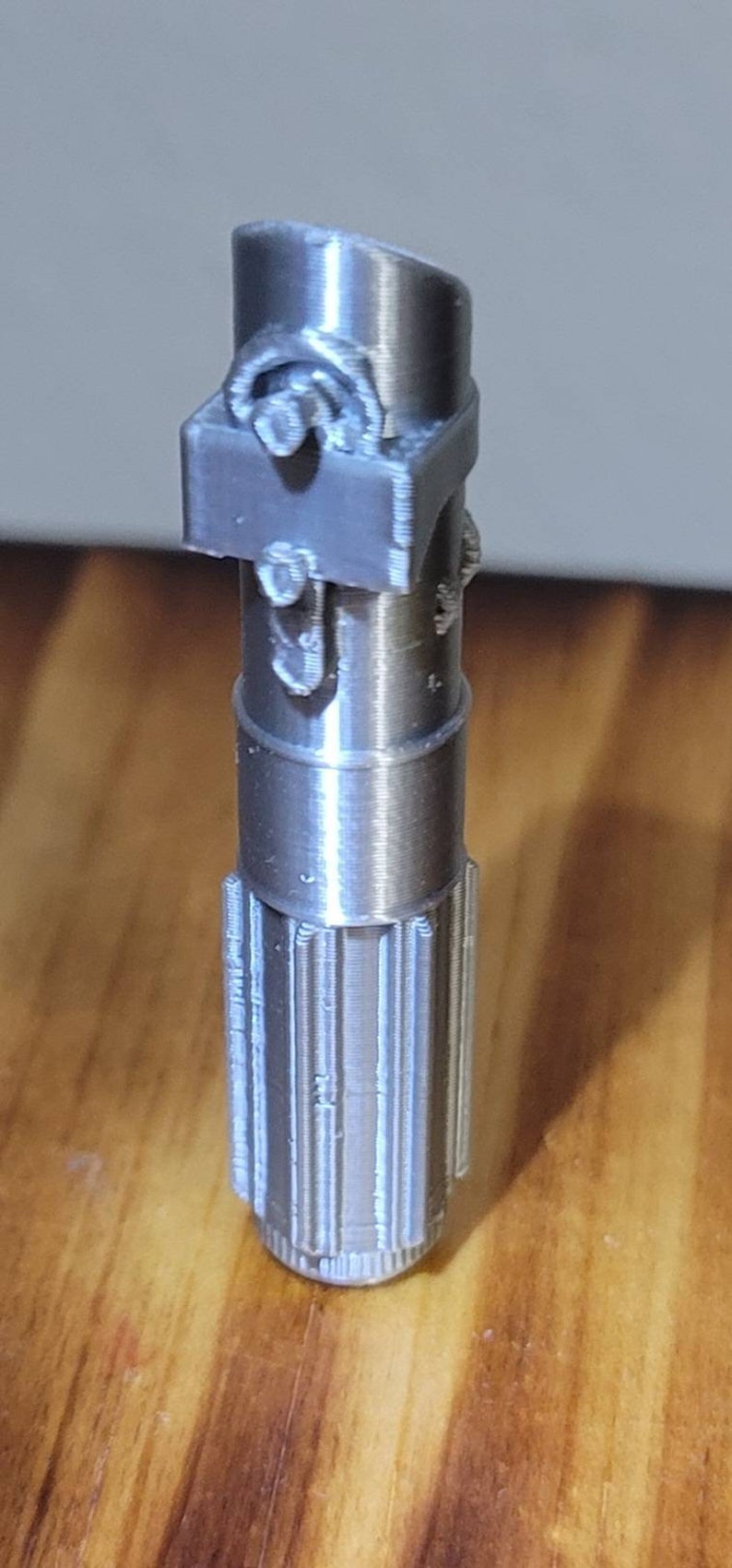 May include: A silver-colored 3D printed lightsaber hilt with a detailed design. The hilt has a textured surface and a cylindrical shape.