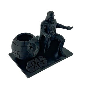 Puede incluir: Un organizador de escritorio negro con temática de Star Wars que presenta a Darth Vader y un modelo de la Estrella de la Muerte. El logotipo de Star Wars se muestra en la base. La figura de Darth Vader está sentada con la mano extendida.