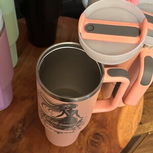 May include: A pink stainless steel travel mug with a black and gray handle and a lid. The mug has a design of a woman in a dress with the word "bridesmaid" on it.