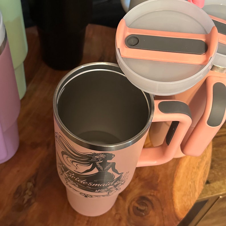 May include: A pink stainless steel travel mug with a black and gray handle and a lid. The mug has a design of a woman in a dress with the word "bridesmaid" on it.