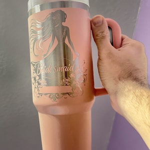 May include: A pink stainless steel travel mug with a handle and a straw. The mug has a silver design of a woman in a dress with the word "bridesmaid" engraved on it.