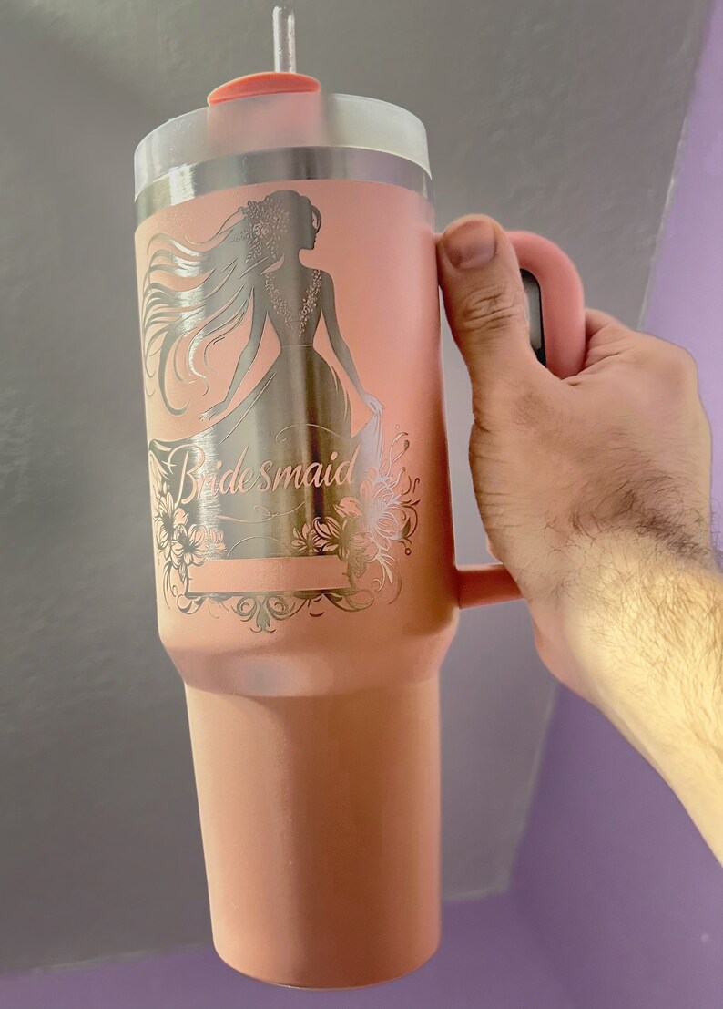 May include: A pink stainless steel travel mug with a handle and a straw. The mug has a silver design of a woman in a dress with the word "bridesmaid" engraved on it.