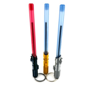 May include: Three lightsaber-shaped pens with keychains. The pens are red, blue, and gold. The keychains are silver, black, and gold.