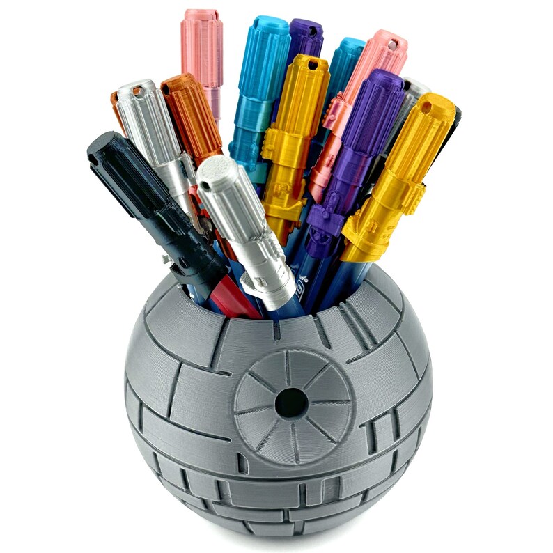 May include: A gray Death Star-shaped pencil holder filled with colorful metallic pens. The pens are arranged in a variety of colors, including pink, blue, green, orange, and purple.