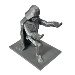 May include: A gray 3D printed figurine of Darth Vader kneeling on a gray base. The figurine is stylized with a low-poly design.