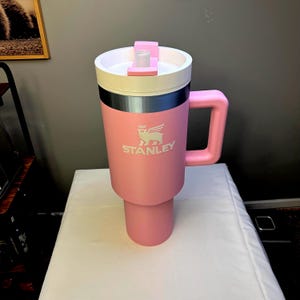 May include: A pink Stanley travel mug with a white lid and a silver band. The mug has a large handle and a white logo that says "STANLEY" with a winged creature above it.