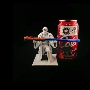 May include: A white plastic figurine of a Star Wars character holding a lightsaber. The figurine is kneeling on a small base. A can of cola with the Star Wars logo and the Mandalorian symbol is in the background.