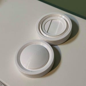 Puede incluir: Dos objetos circulares blancos impresos en 3D. Uno está abierto, revelando un interior empotrado con un disco central y un canal circundante. El otro está cerrado, con un diseño de círculos concéntricos texturizados en su superficie superior. Son probablemente tapas.
