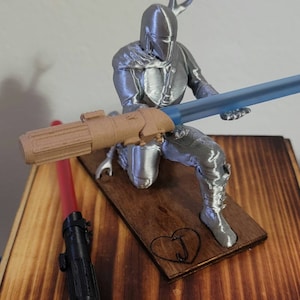 May include: A silver 3D printed figurine of a Mandalorian character holding a blue lightsaber. The figurine is standing on a wooden base with a heart and the letter "J" carved into it. A red and black lightsaber is lying on the wooden surface.