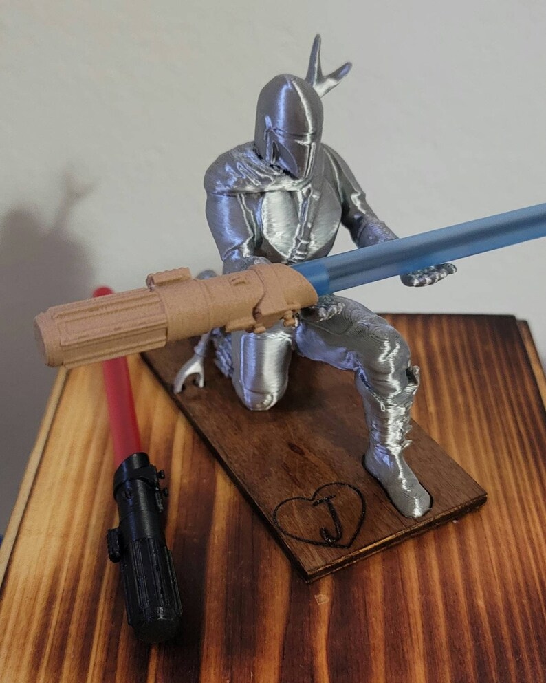 May include: A silver 3D printed figurine of a Mandalorian character holding a blue lightsaber. The figurine is standing on a wooden base with a heart and the letter "J" carved into it. A red and black lightsaber is lying on the wooden surface.