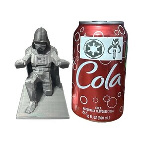 May include: A 3D printed, silver-colored figurine of Darth Vader kneeling on a square base. The figurine is next to a red can of cola with a white label that says "Cola" and has a white star wars logo and a Mandalorian symbol.