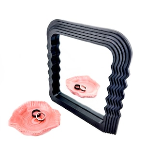 May include: A black, wavy framed mirror reflects a pink dish with two silver rings. The dish is sitting in front of the mirror on a white surface.