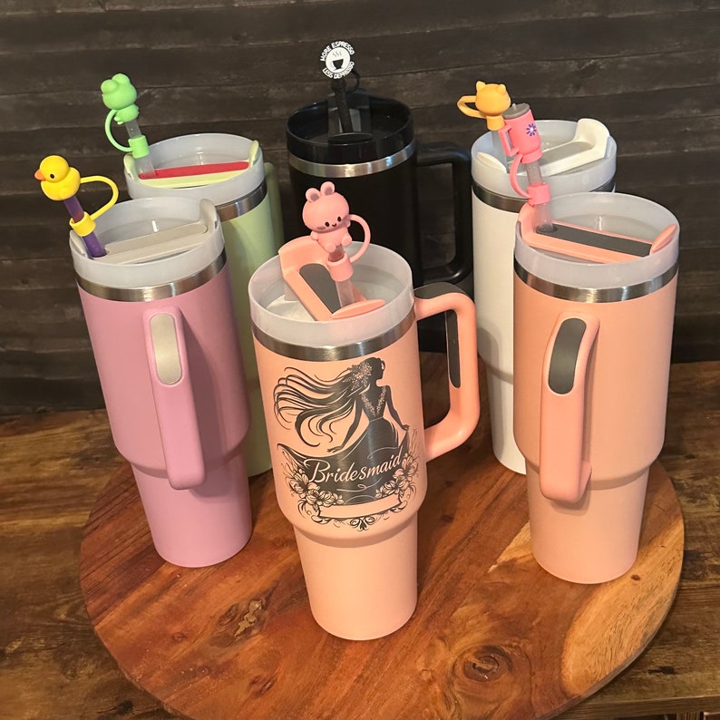 May include: Six stainless steel travel mugs with various colored lids and straws. The mugs are in different colors, including pink, green, white, and black. One mug has the word "Bridemaid" printed on it.