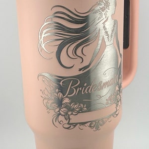 May include: A pink stainless steel tumbler with a silver lid and handle. The tumbler has a laser engraved design of a bride with the word "Bridesmaid" on it.