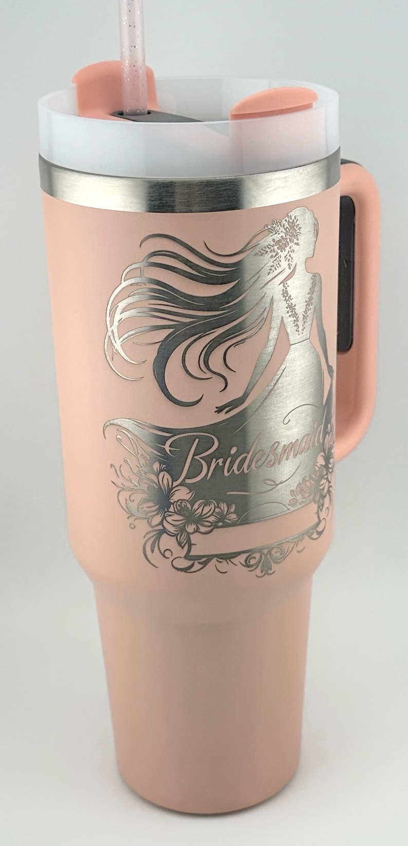 May include: A pink stainless steel tumbler with a silver lid and handle. The tumbler has a laser engraved design of a bride with the word "Bridesmaid" on it.