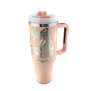May include: A pink stainless steel travel mug with a black handle. The mug has a silver design of a woman with the words "Bridemaid Valentine" on it.