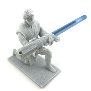 May include: A gray plastic figurine of a Star Wars character holding a blue lightsaber. The character is kneeling on a gray base.