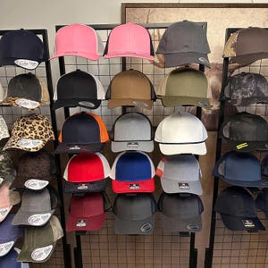 May include: A display of various trucker hats in a variety of colors, including pink, black, gray, and brown. Some hats have mesh backs and rope detailing. The hats are arranged on a black metal grid.