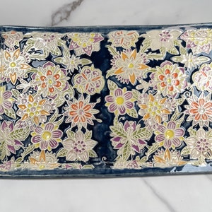 May include: A rectangular ceramic serving tray with a dark blue background and a raised floral design in shades of white, yellow, orange, and purple.