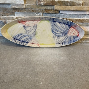 May include: A white ceramic oval serving dish with a colorful abstract design. The design features blue, yellow, red, and white swirls and lines.