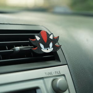 Op de afbeelding: Een zwarte, rode en witte ventilatieclip met het personage Shadow the Hedgehog. De clip is bevestigd aan een ventilatierooster van een auto, met de radioknoppen eronder zichtbaar. De tekst "TUNE • FILE" staat op de radio.
