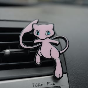 May include: A pink cartoon character air freshener with black outlines, featuring blue eyes and a long tail. The air freshener is attached to a car vent. The car's dashboard has the text "TUNE FILE".