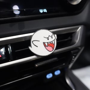 May include: A white, circular air vent charm featuring a cartoon ghost with a wide, toothy grin and a pink tongue. The charm is placed on a car's air vent, with the dashboard and controls visible in the background.