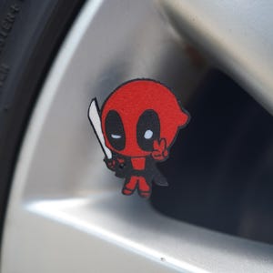 Op de afbeelding: Een rode en zwarte cartoon karakter sticker met een wit zwaard en vredesteken handgebaar. De sticker is bevestigd aan een zilveren autowiel. Het karakter heeft grote zwarte ogen en een rode kop.