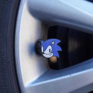 May include: A blue and white Sonic the Hedgehog valve stem cap. The cap is attached to a silver car wheel.
