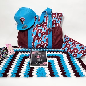 May include: A set of blue and maroon themed items including a tote bag, a baseball cap, a small pouch, a crocheted blanket, and a journal with the words "Brave Hope Strong".