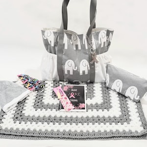 May include: A gray and white tote bag with a pattern of elephants, a gray and white crocheted blanket, a gray and white pencil case, a gray beanie, a pink and white journal with the text "Brave Hope Strong", and a pink and white hair clip.