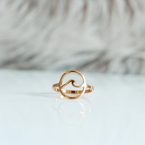 Puede incluir: Un anillo de oro con un diseño de ola. El anillo está sobre una superficie blanca.