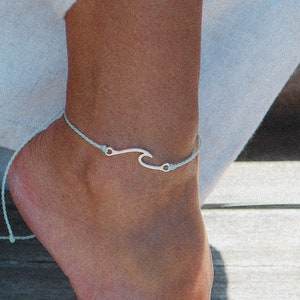 May include: A light blue ankle bracelet with a silver wave charm. The bracelet is tied on a person's ankle.
