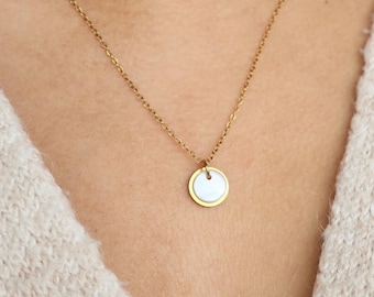 May include: A gold-colored necklace with a small, round pendant. The pendant has a white center and a gold-colored border. The necklace is worn against a light beige sweater.