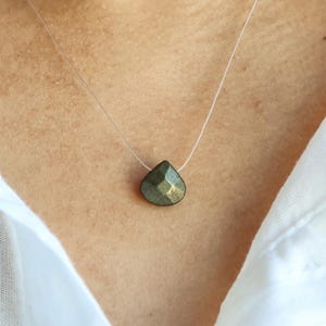 May include: A teardrop-shaped pyrite pendant necklace. The pendant is a dark, metallic green with gold flecks, suspended on a nearly invisible clear cord. The necklace is worn against a white shirt.