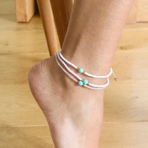 May include: Delicate anklets with white, pink, and clear beads, accented with turquoise beads. The anklets are layered, creating a subtle, elegant look. The jewelry is worn on the ankle.