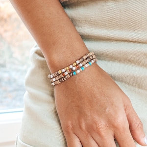 May include: Three beaded bracelets with a mix of neutral and colorful beads are stacked on a wrist. The bracelets feature brown, white, yellow, red, and turquoise beads. The person is wearing light-colored shorts.