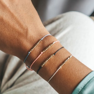 Puede incluir: Una colección de cinco pulseras minimalistas. Cada pulsera presenta un cordón fino en varios colores: azul claro, rosa, negro y blanco. Los cordones están adornados con una fila de pequeñas cuentas doradas.